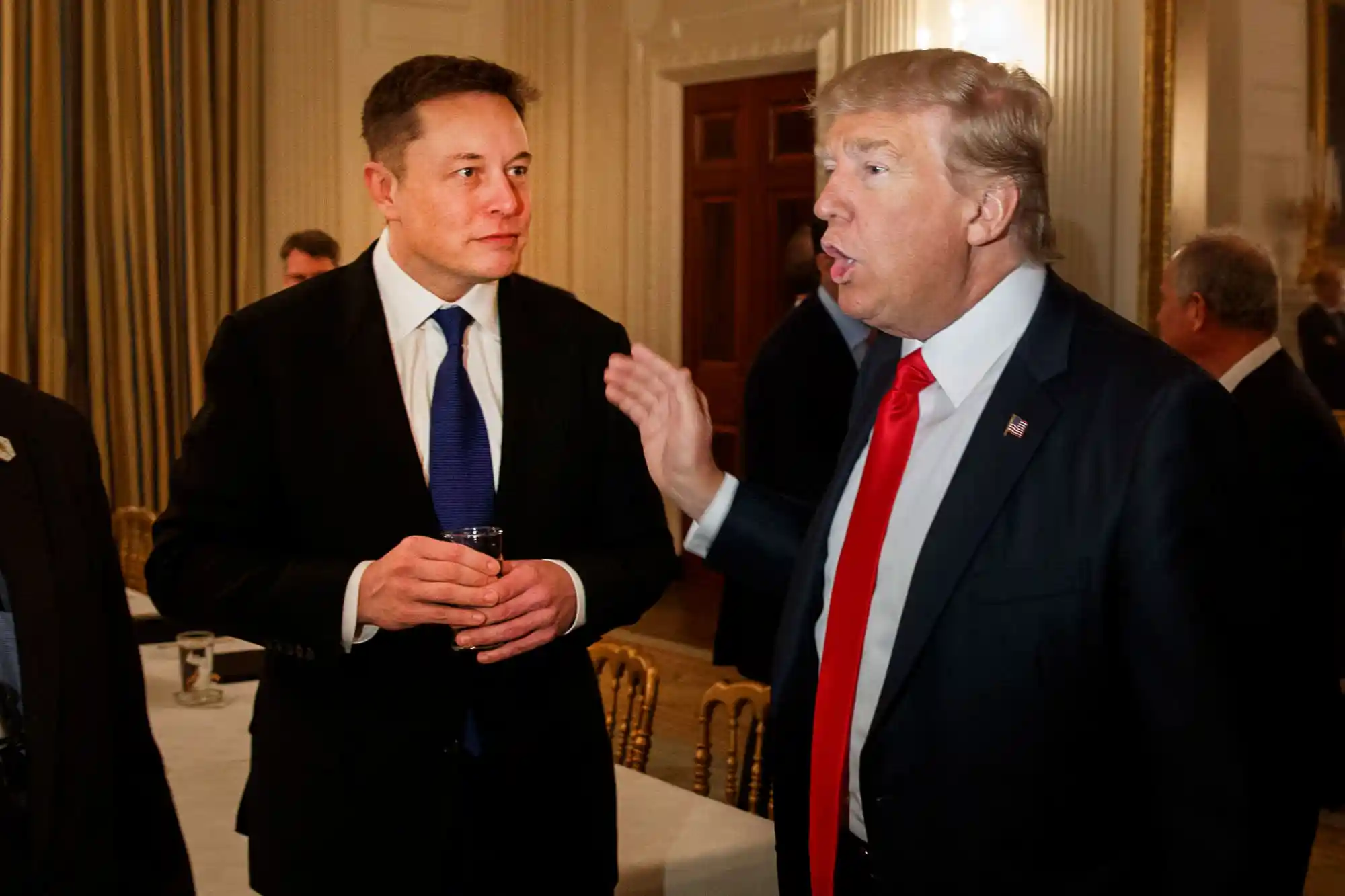 The image shows a man with a serious and expressive face, with blond hair combed to the side and dressed in a dark suit, white shirt, and red tie. His gaze is direct, and his expression conveys determination or concentration. The background is slightly blurred, helping to focus attention on the face and details of the main figure. The photograph appears to be taken in a formal or professional setting, possibly in a government building or high-profile context.